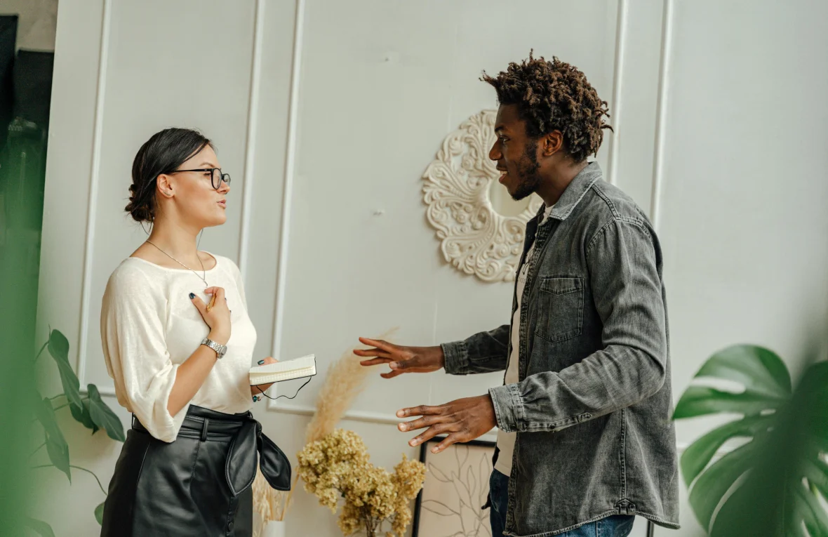 A man and woman standing and talking in a bright room, with the woman holding a notebook and the man gesturing, symbolizing Dialectical Behavioral Therapy (DBT) in Tempe AZ.